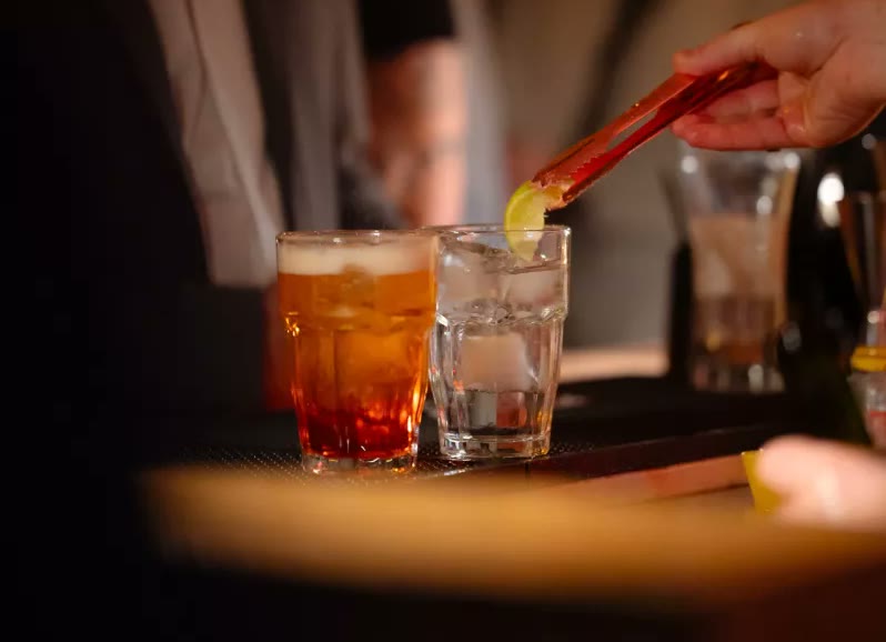 Le mani del barista lasciano cadere una fetta di limone nel bicchiere, accanto a un cocktail rosso sul bancone